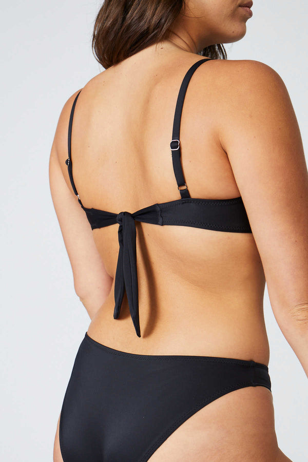 Woman wearing a Bambina Swim black two piece bikini, ring detail on strap, tie back, mid coverage bottom, high cut leg.
