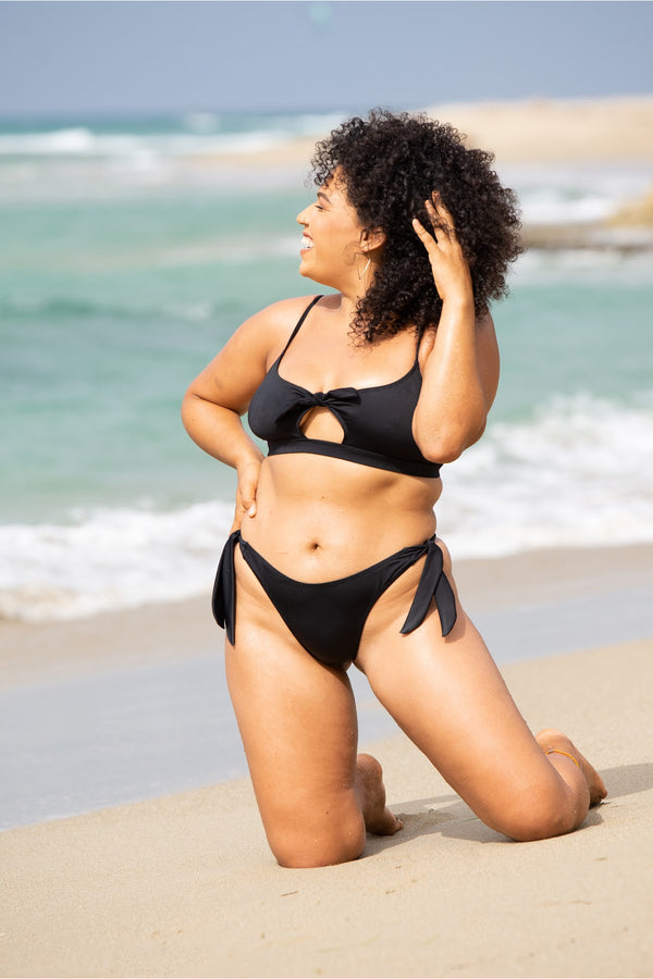 Serena Thong Bikini Bottom in black, front view on model on beach, showing high cut leg and side ties.