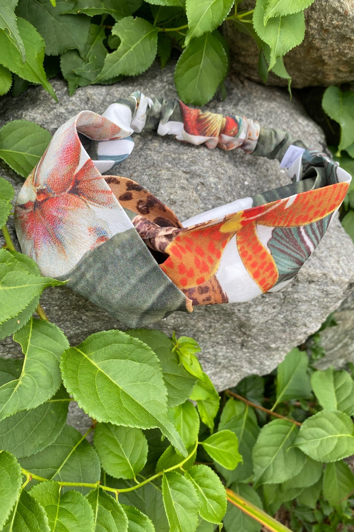 Bambina Swim Cardamom headband in tropical print, placed on a rock with greenery, flat lay image showing overall shape and tropical pattern.
